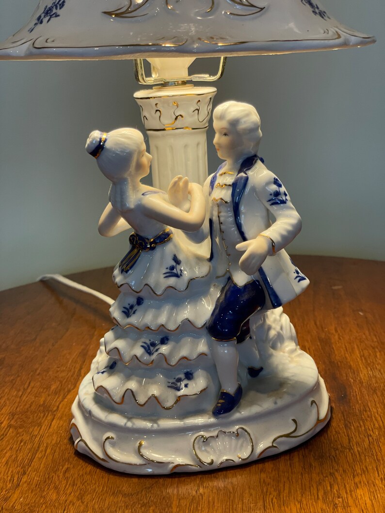 May include: A white porcelain table lamp with a blue and white floral design. The lamp features a couple dancing, with the woman in a white dress and the man in a blue and white suit.