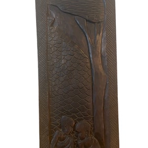 May include: A dark brown wooden wall hanging with a carved design of three people sitting under a tree. The background is a textured pattern of scales.