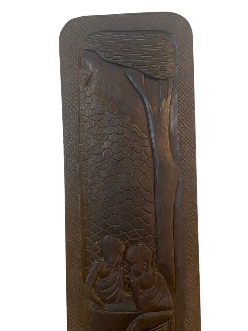 May include: A dark brown wooden wall hanging with a carved design of three people sitting under a tree. The background is a textured pattern of scales.