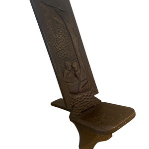 May include: A dark brown wooden folding chair with intricate carvings of people and a tree. The chair has a simple design with a flat seat and a back that folds down.