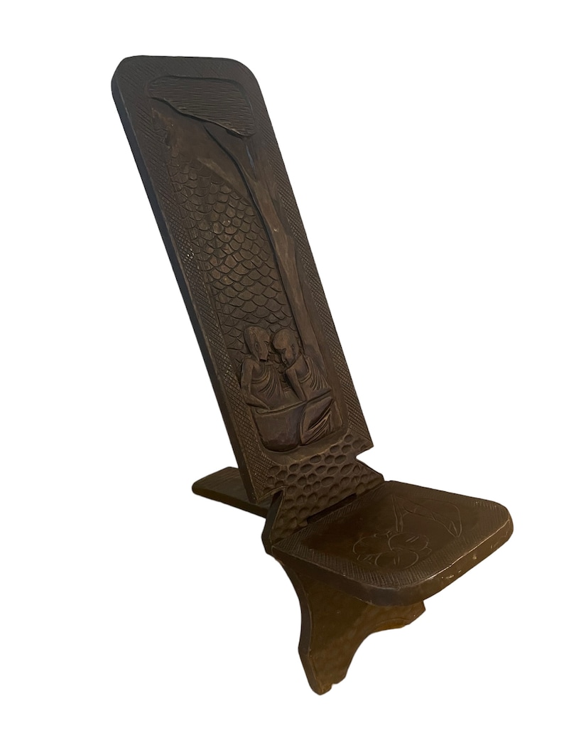 May include: A dark brown wooden folding chair with intricate carvings of people and a tree. The chair has a simple design with a flat seat and a back that folds down.