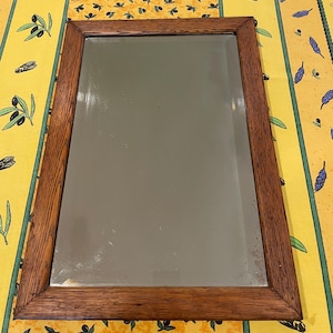 May include: A rectangular wall mirror featuring a medium-brown wood frame with a visible grain pattern. The glass surface is clear and reflective, showing minor signs of age and wear along the edges. The frame has mitered corners.
