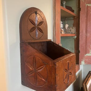 May include: A vintage wooden wall-mounted box with a hinged lid. The box is stained a warm brown and features carved starburst designs on the front, sides, and lid. The box is empty and mounted on a light-colored wall.