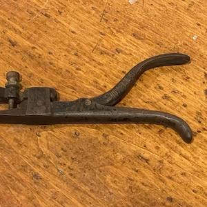 May include: A vintage saw set tool, made of dark metal, rests on a wooden surface. The tool features a lever handle and adjustable components, likely used for setting the teeth of a saw blade. The tool's design suggests it is an antique or a well-used item.