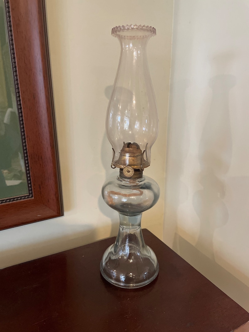 May include: A clear glass oil lamp with a frosted glass chimney and a brass burner. The lamp has a round base and a decorative, scalloped rim.