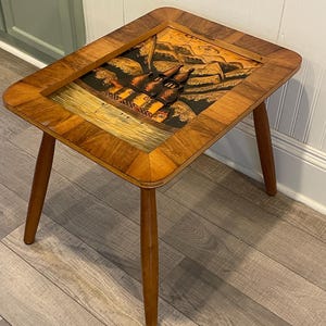 May include: A vintage wooden side table with a rectangular top and three tapered legs. The tabletop features a framed, decorative panel with a landscape scene in shades of brown, orange, and black. The table is made of wood with a warm, natural finish.