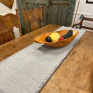 Puede incluir: Una mesa de madera con un camino de mesa tejido azul y blanco. Un cuenco de madera con una calabaza amarilla, un calabacín verde, un pimiento rojo y un aguacate está sobre el camino de mesa.