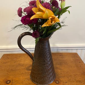 Op de afbeelding: Een boeket roze en gele bloemen in een bruine, getextureerde koperen kan.