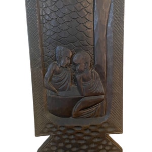 May include: A dark brown wooden chair with a carved design of three figures. The chair has a high back and a seat with a carved pattern.