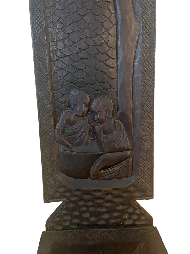 May include: A dark brown wooden chair with a carved design of three figures. The chair has a high back and a seat with a carved pattern.