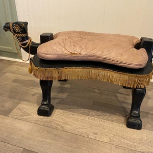 May include: A black wooden camel-shaped footstool featuring a pinkish-tan leather cushion on its back. The stool has gold-colored fringe trim along the base and a decorative gold rope harness around the camel's neck, with four sturdy legs.