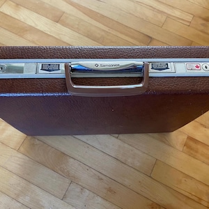 Pode incluir: Maleta Samsonite castanha com exterior texturizado. A mala tem uma estrutura e pega prateadas, com o logótipo Samsonite visível. A mala tem fechadura de combinação e pega para transporte.