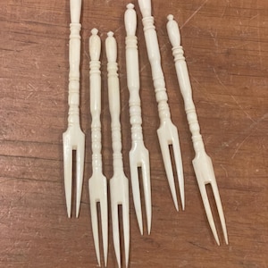 Può includere: Una collezione di sei forchette da dessert antiche color avorio. Ogni forchetta presenta un manico decorativo con una parte superiore arrotondata e una forchetta a due rebbi. Le forchette sono disposte su una superficie di legno.