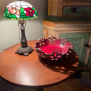 May include: A stained glass table lamp with a floral design in red, pink, and green sits on a round wooden table. A decorative red bowl with a cut-out floral pattern is also on the table. The lamp has a dark metal base.