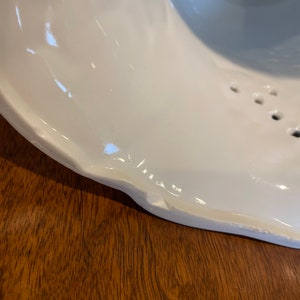 May include: White ceramic bowl with a chipped edge. The bowl has a textured surface and a series of small holes around the rim.