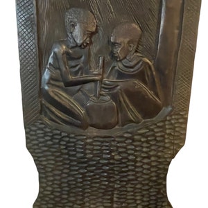 May include: A dark brown wooden chair with a carved design of two people sitting and stirring a pot. The chair has a woven pattern on the back and bottom.