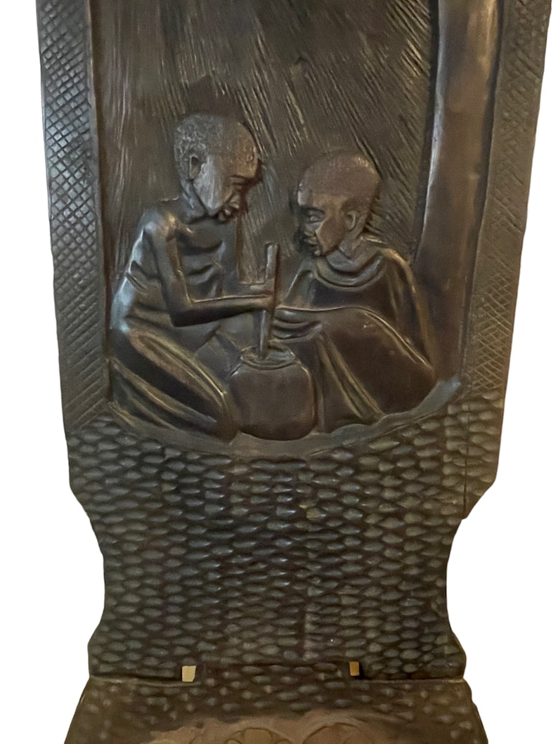 May include: A dark brown wooden chair with a carved design of two people sitting and stirring a pot. The chair has a woven pattern on the back and bottom.