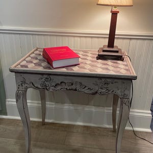 May include: A small, antique-style side table with a checkered top and ornate, curved legs. A red book rests on the table, next to a lamp with a brown base and beige shade. The table is painted in a light, neutral color.