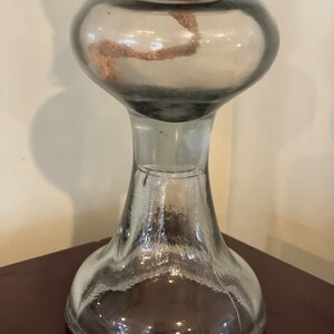 May include: A clear glass oil lamp with a round, bulbous top and a wide, flared base. The lamp has a metal wick holder and a metal cap on top.