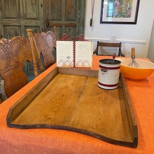 May include: A rectangular wooden dough board with raised sides, a white and red metal flour sifter with a crank handle, a red wire cookbook stand holding an open recipe book, and a yellow mixing bowl with a wire whisk inside on an orange cloth.