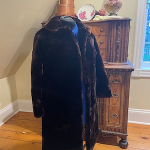May include: A long brown faux fur coat with a button closure. The coat is on a mannequin and is displayed against a wooden background.