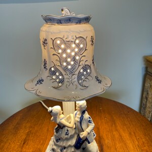 May include: A white porcelain table lamp with a blue and white floral design. The lampshade has a cut-out design and is illuminated by a light bulb. The base of the lamp features a couple dancing.