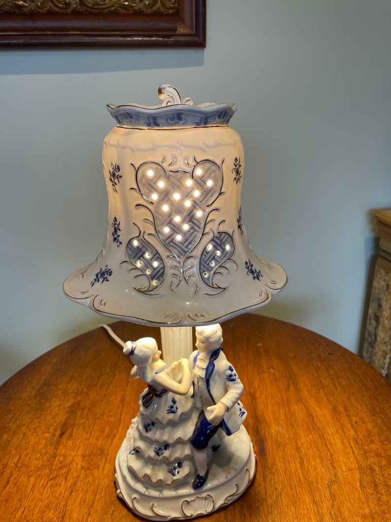 May include: A white porcelain table lamp with a blue and white floral design. The lampshade has a cut-out design and is illuminated by a light bulb. The base of the lamp features a couple dancing.