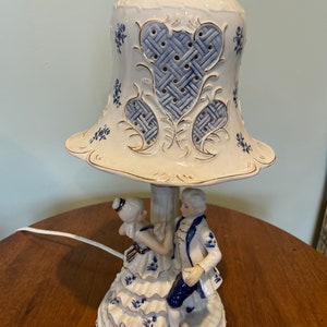May include: A porcelain table lamp with a blue and white design. The lamp features a couple dancing on the base and a bell-shaped shade with a lattice pattern.