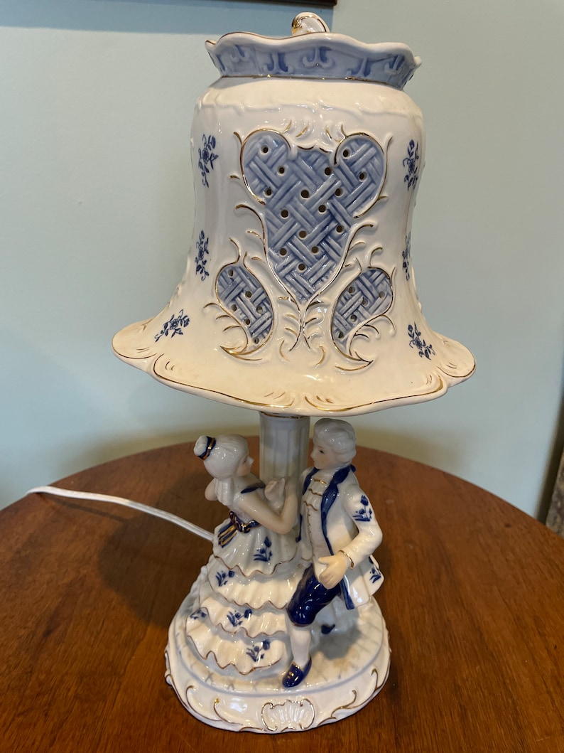 May include: A porcelain table lamp with a blue and white design. The lamp features a couple dancing on the base and a bell-shaped shade with a lattice pattern.