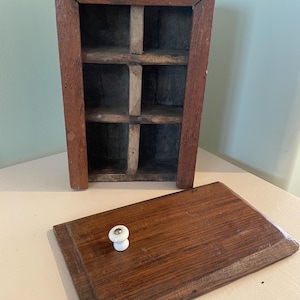 May include: A rectangular wooden wall-mounted storage box with a dark brown finish and six internal compartments. The box features a removable wooden lid with a small, round white ceramic knob attached to the center for easy opening and closing.