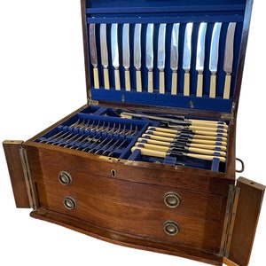 May include: A wooden cutlery box with two drawers and a hinged lid. The lid is open and reveals a blue velvet lining and a set of silver knives with ivory handles.