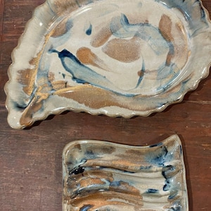 May include: Two ceramic serving dishes with a blue and brown glaze. The larger dish is oval shaped with a scalloped edge. The smaller dish is rectangular with a wavy edge.