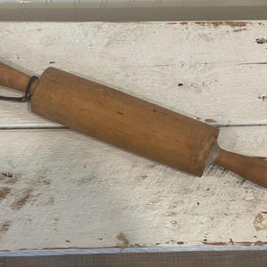 May include: A wooden rolling pin with a cylindrical body and two rounded handles. The rolling pin is light brown and has a leather loop attached to one end. It rests on a white, distressed wooden surface.
