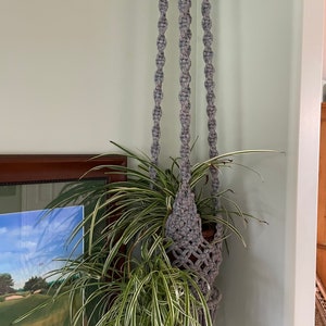 Peut inclure: Un porte-plante en macramé gris avec des perles en bois suspendu au plafond et tenant une plante en pot avec de longues feuilles vertes.