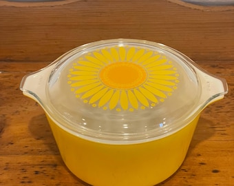 May include: A yellow Pyrex casserole dish with a clear lid featuring a yellow and orange sunflower design. The dish has two handles and is likely vintage. The dish is a bright yellow color.