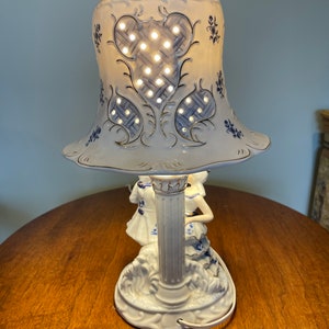 May include: A white porcelain table lamp with a blue and gold floral design. The lamp features two figures of women in blue and white dresses, and a bell-shaped shade with a lattice pattern.