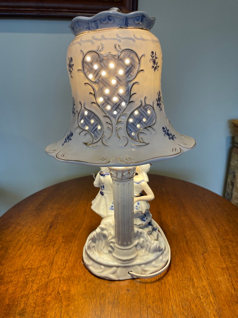 May include: A white porcelain table lamp with a blue and gold floral design. The lamp features two figures of women in blue and white dresses, and a bell-shaped shade with a lattice pattern.