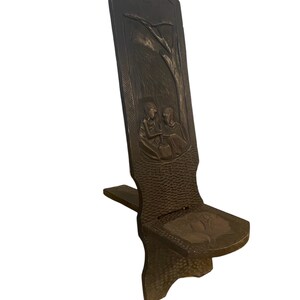 May include: A dark brown wooden chair with a carved design of two people sitting under a tree. The chair has a simple, rustic design and is made of solid wood.