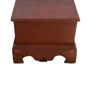 May include: A red wooden storage chest with a hinged lid and a base with carved details. The chest is painted a deep red color and has a rustic appearance.
