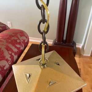 May include: A gold-colored metal ceiling light fixture with a square base and a chain hanging from it. The chain is made of metal and has a yellow wire running through it.