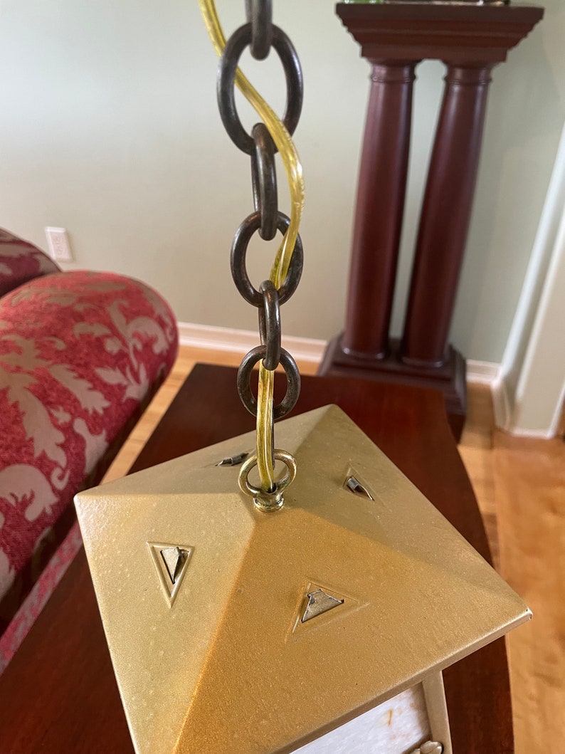 May include: A gold-colored metal ceiling light fixture with a square base and a chain hanging from it. The chain is made of metal and has a yellow wire running through it.