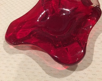May include: A red glass ashtray with a wavy, organic shape. The ashtray is made of clear glass with a red tint.