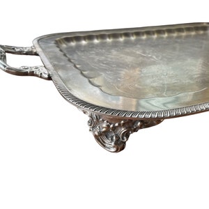 May include: A silver serving tray with ornate handles and feet. The tray has a slightly raised edge and a smooth, reflective surface.