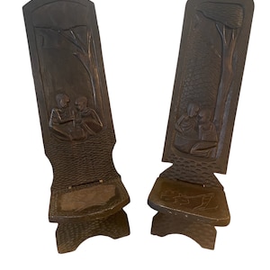 May include: Two dark brown wooden folding chairs with carved figures and trees. The chairs have a textured surface and are made of a single piece of wood.