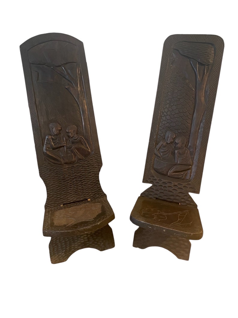 May include: Two dark brown wooden folding chairs with carved figures and trees. The chairs have a textured surface and are made of a single piece of wood.