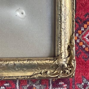 May include: An ornate gold-colored picture frame featuring intricate relief carvings along the edges and corners. The frame surrounds a plain, light gray mat board with a small, circular white reflection visible on the surface of the matting.