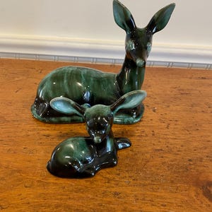 May include: A pair of ceramic deer figurines in a green and black glaze. The larger deer is sitting with its head up, while the smaller deer is lying down.