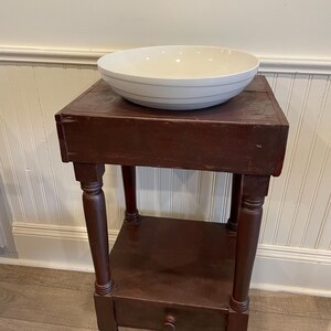 May include: A dark red wooden table with a single drawer and two shelves. A white ceramic bowl sits on the top shelf.