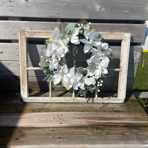 May include: A white magnolia wreath with green leaves and white berries, displayed in a vintage white window frame.