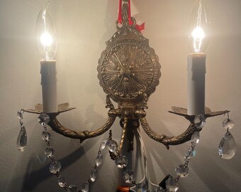 May include: Vintage brass wall sconce with two candle-shaped light bulbs. The sconce features ornate detailing, crystal accents, and a decorative medallion. The sconce is mounted on a wall with a red ribbon.